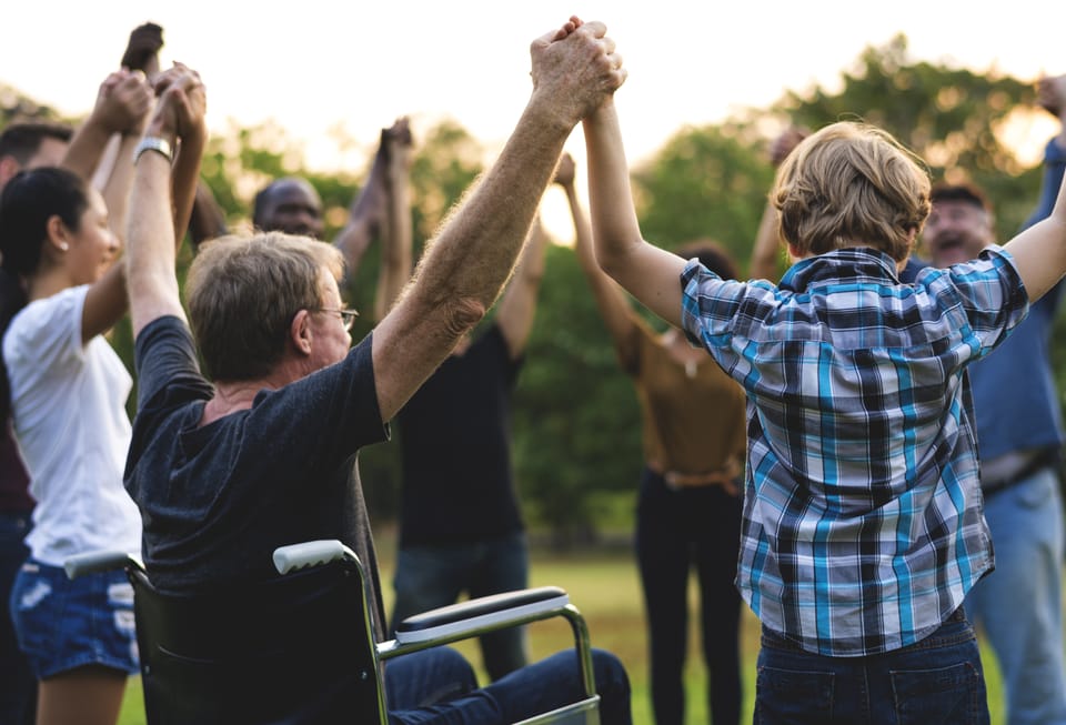 A group of people, including a wheelchair user, raising their joined hands together outdoors